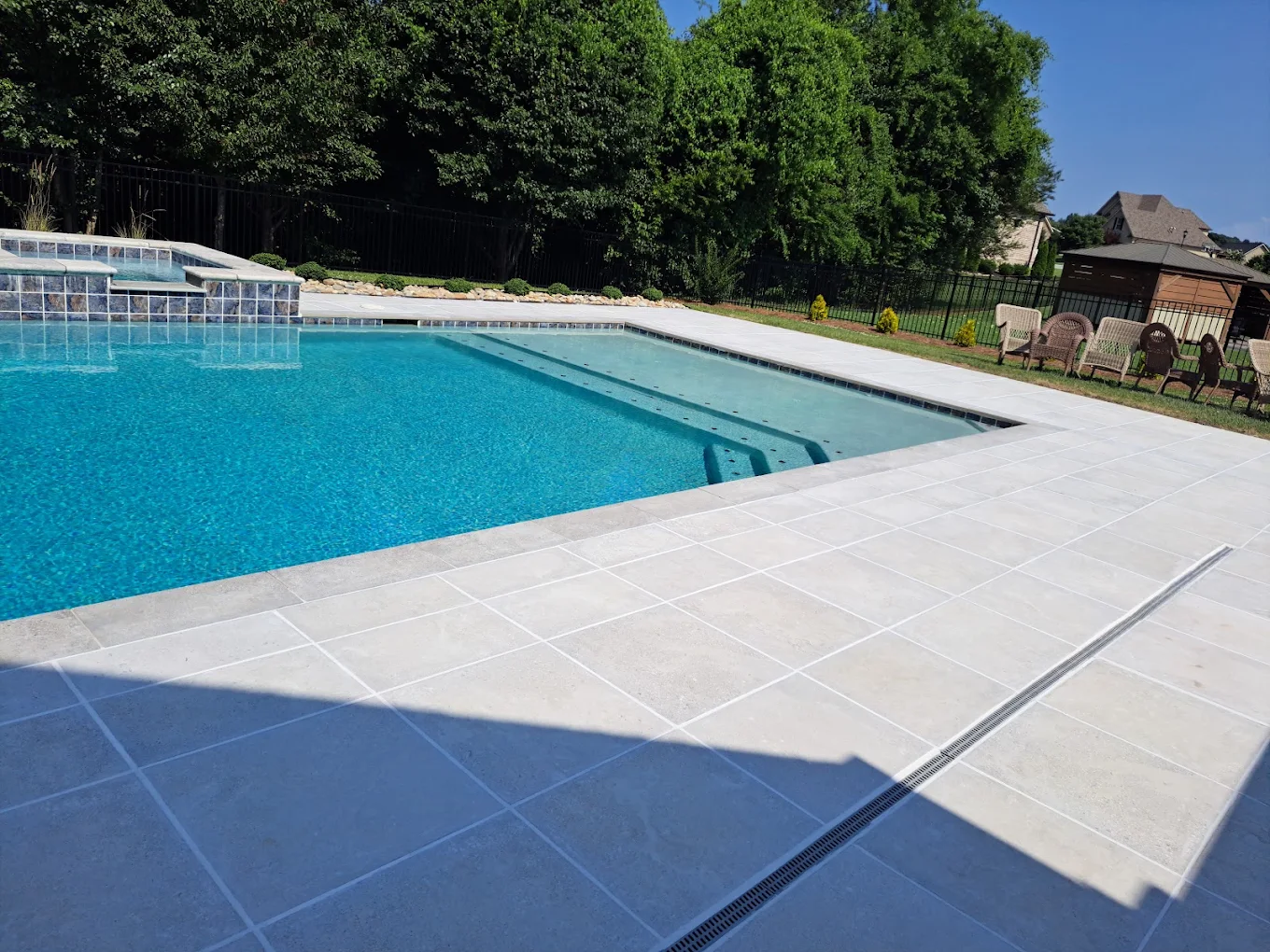 Residential swimming pool with clear blue water, white concrete deck, lounge chairs, and evergreen trees in background
