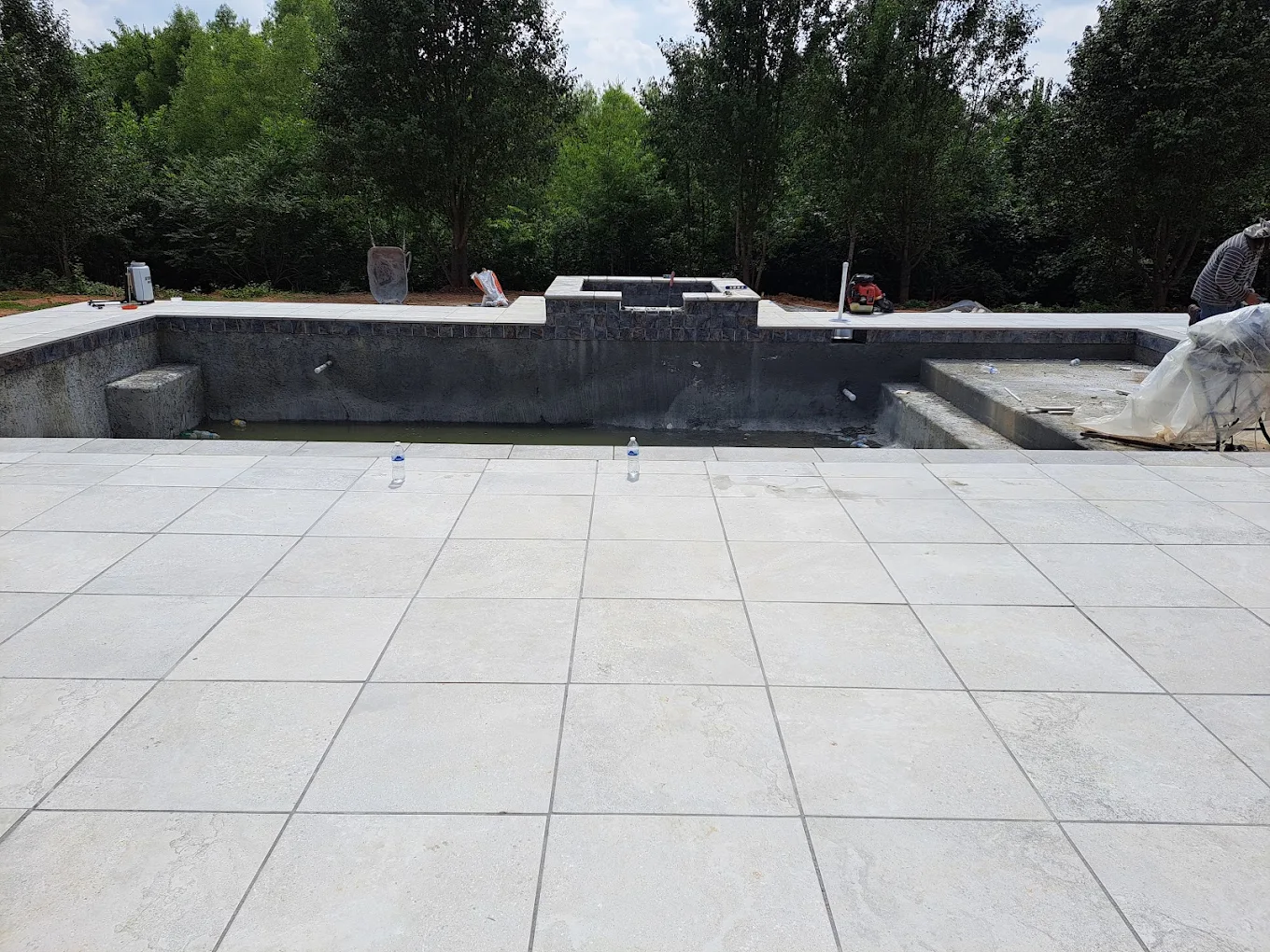 Empty concrete pool under construction with paved deck and wooded backdrop