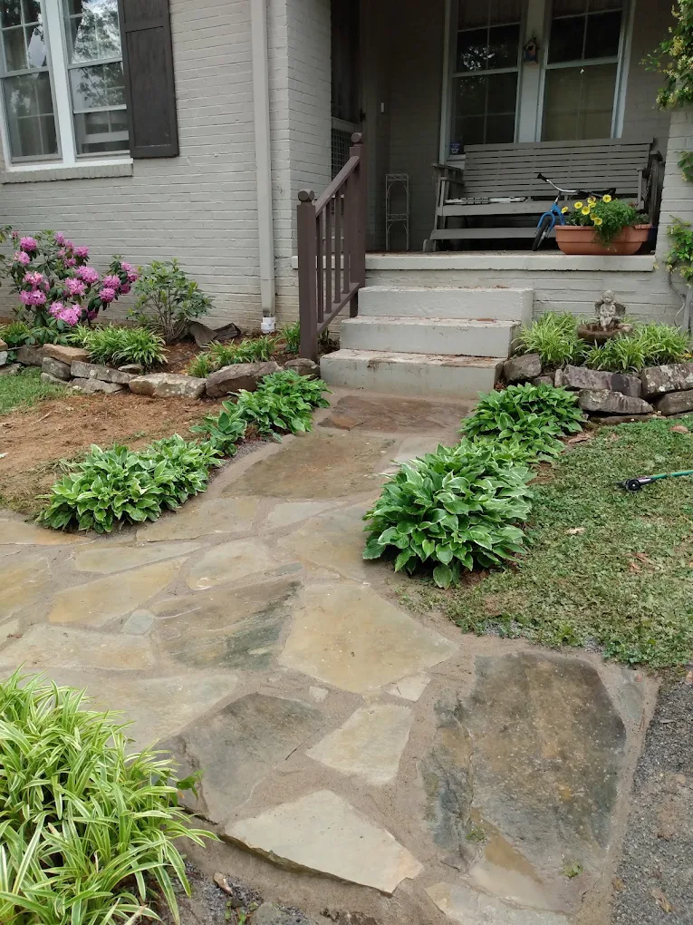 Front entrance of brick house with stone pathway, steps, pink flowers, potted plants, and wooden porch railing