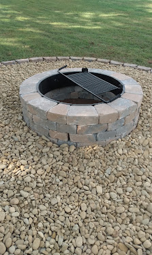 Circular stone fire pit with black metal grate on gravel base with grass background