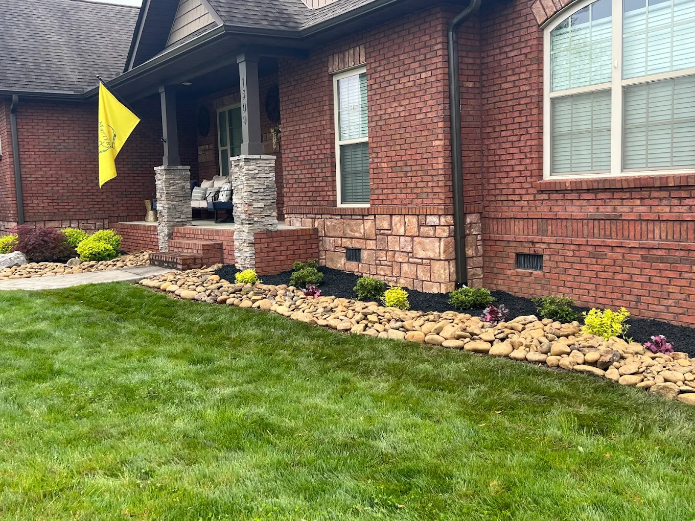 Brick home with stone porch pillars, yellow flag, landscaped rock garden with shrubs and mulch