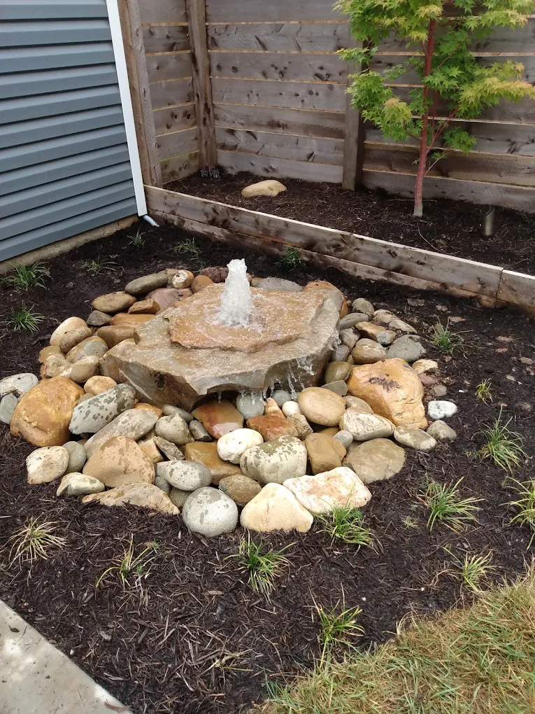 Small water fountain surrounded by river rocks in dark mulch garden bed with wooden fence and climbing plants
